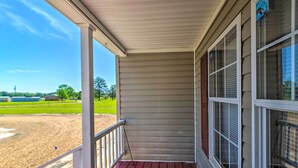 A covered porch with a view of a sandy field and green grass.