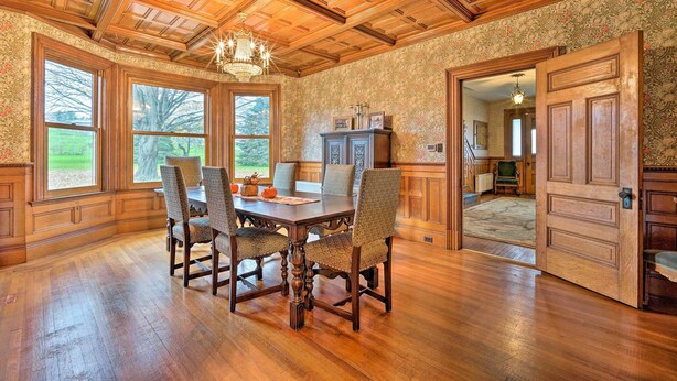 A dining room with a wooden table and chairs, large windows, and a chandelier.
