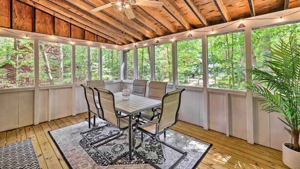 Eine überdachte Terrasse mit Holzboden, einem Deckenventilator, einem Tisch mit Stühlen und großen Fenstern mit Blick auf den umliegenden Wald.