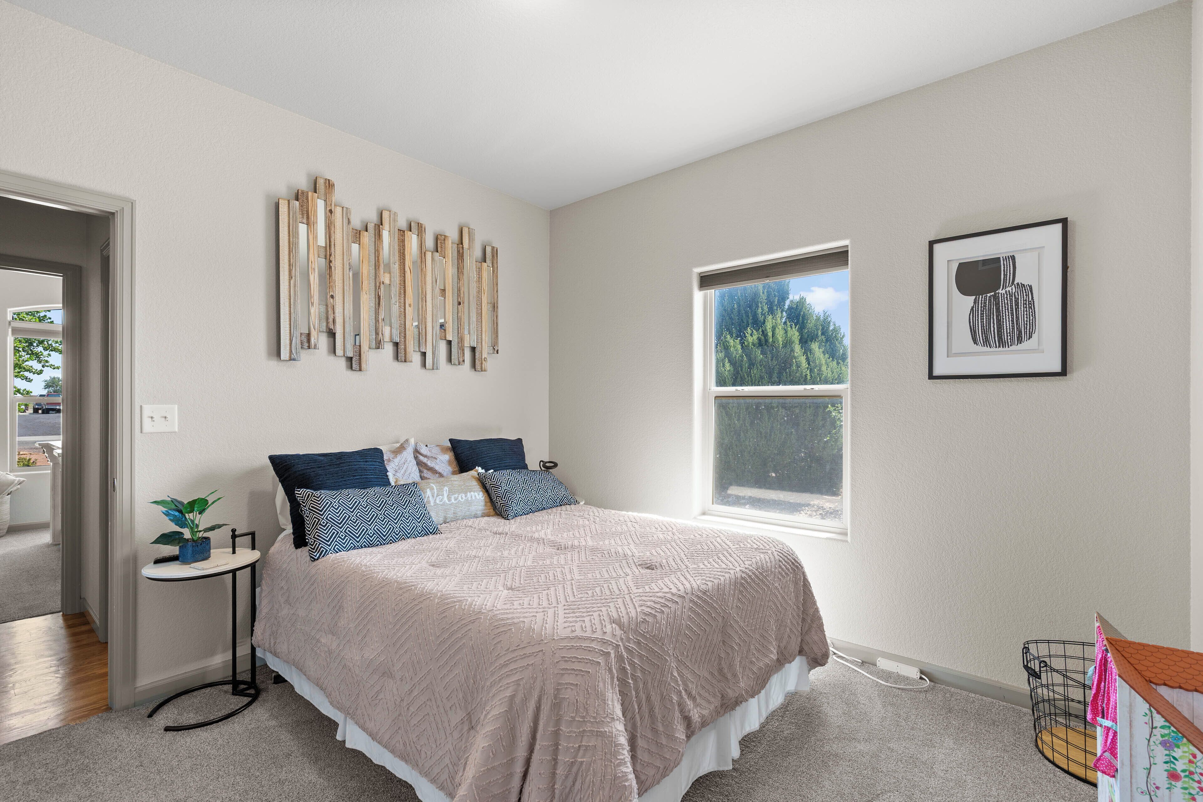 A bedroom with a bed, a window, a wall-mounted artwork, and a small side table with a plant.