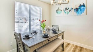 A dining area with a table set for four, a bench, and a window with blinds. There are three wall-mounted artworks and a chandelier above the table. - Garden City Vacation Rental - 1 Mi to Beach! (Garden City)