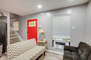Ein Zimmer mit einem sofa in Zickzack-Muster, einem dunkelbraunen Sofa, einer roten Tür, einem kleinen Tisch mit einer Lampe und einem Fenster mit Jalousien.