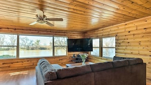 Interieur van een blokhut met een groot houten plafond, een bank, een flatscreen-televisie en een raam met uitzicht op een natuurlandschap.