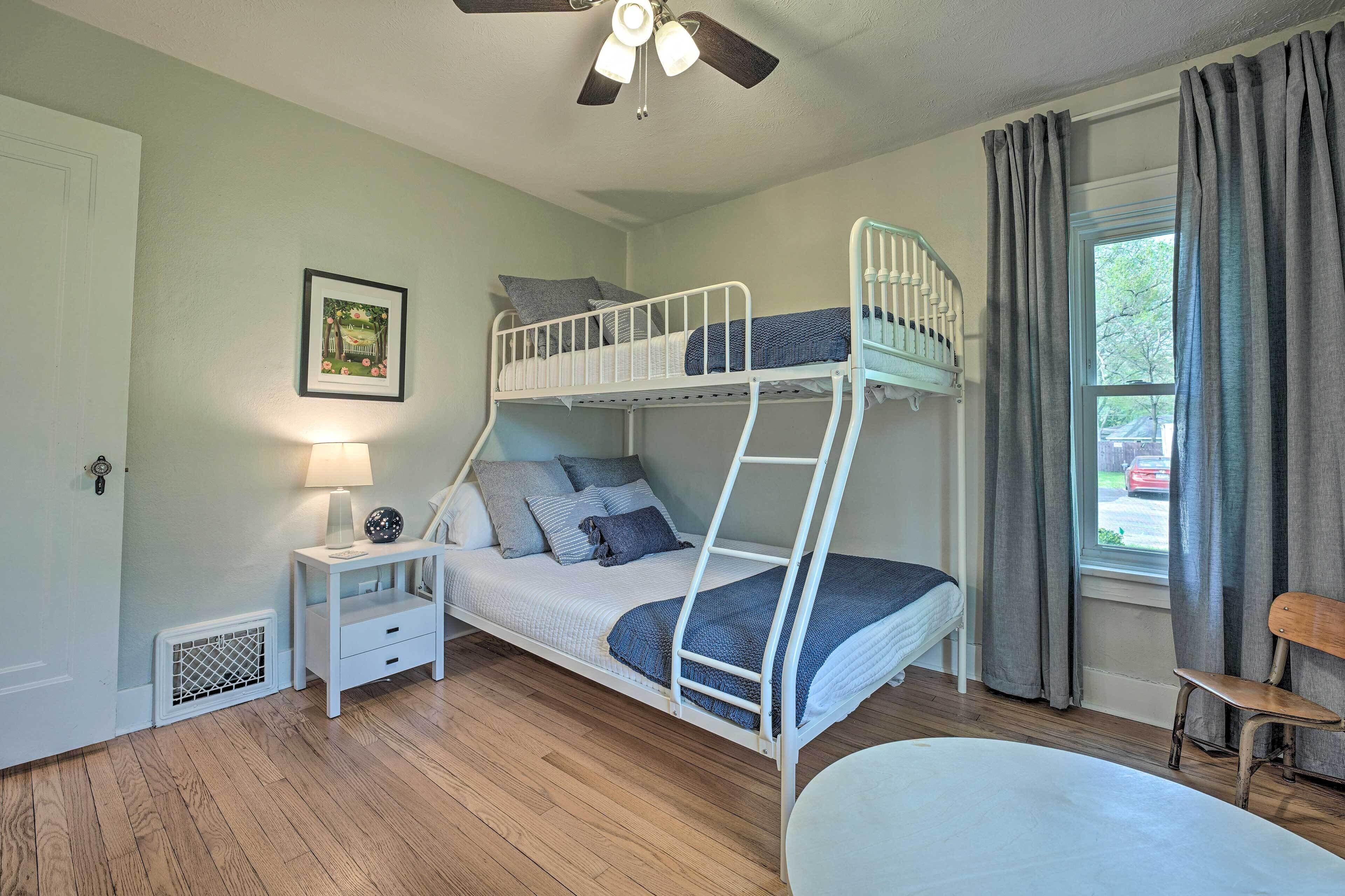 A bunk bed room with a ceiling fan, a nightstand with a lamp, a window with curtains, and a view of the outside.