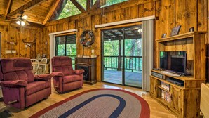 Interior de una cabaña de madera con dos sillones rojos, una chimenea, un televisor sobre un mueble de madera y un ventanal con vistas a una terraza.