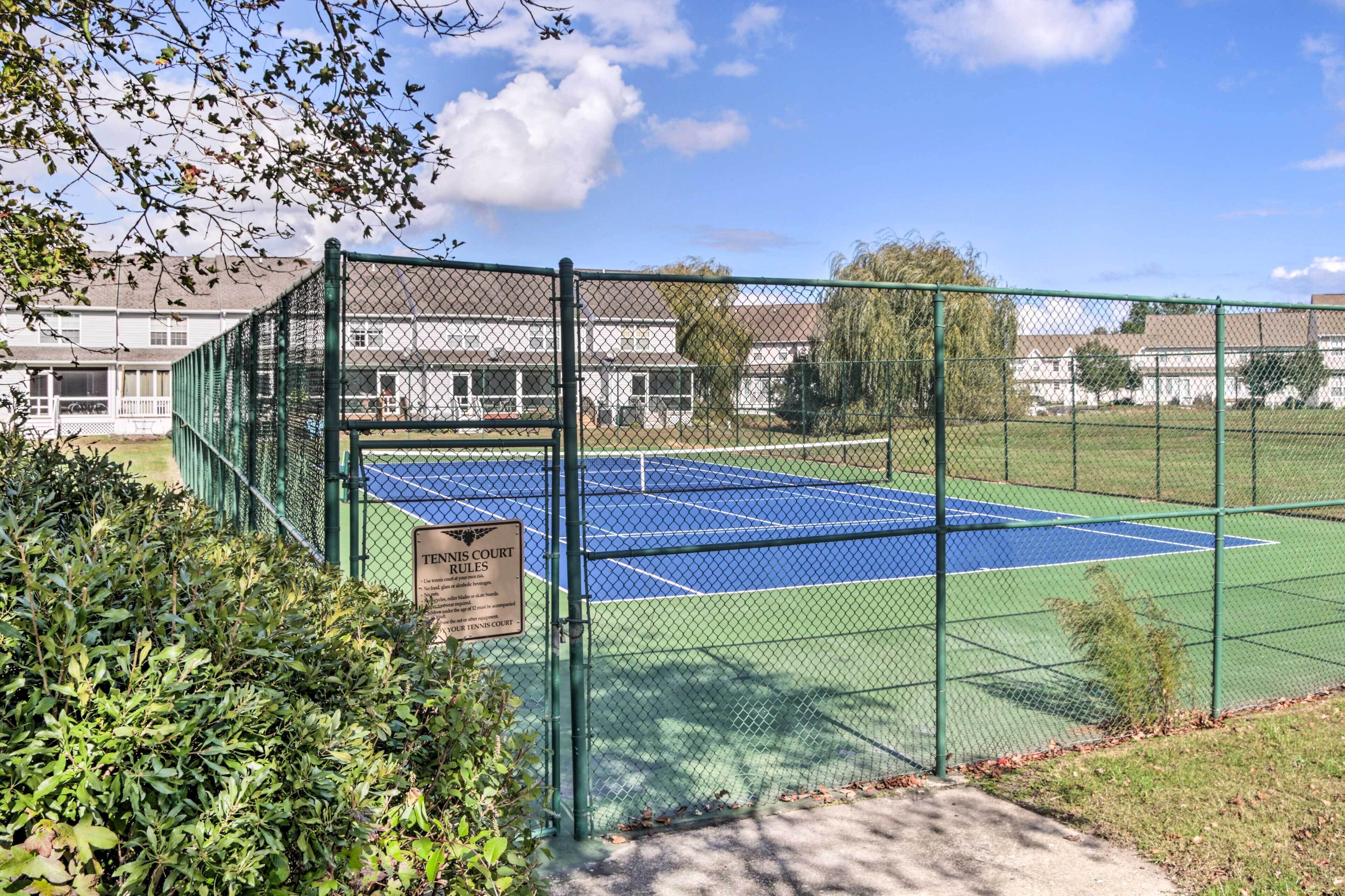 A tennis court enclosed by a green fence, surrounded by a grassy area and residential buildings.