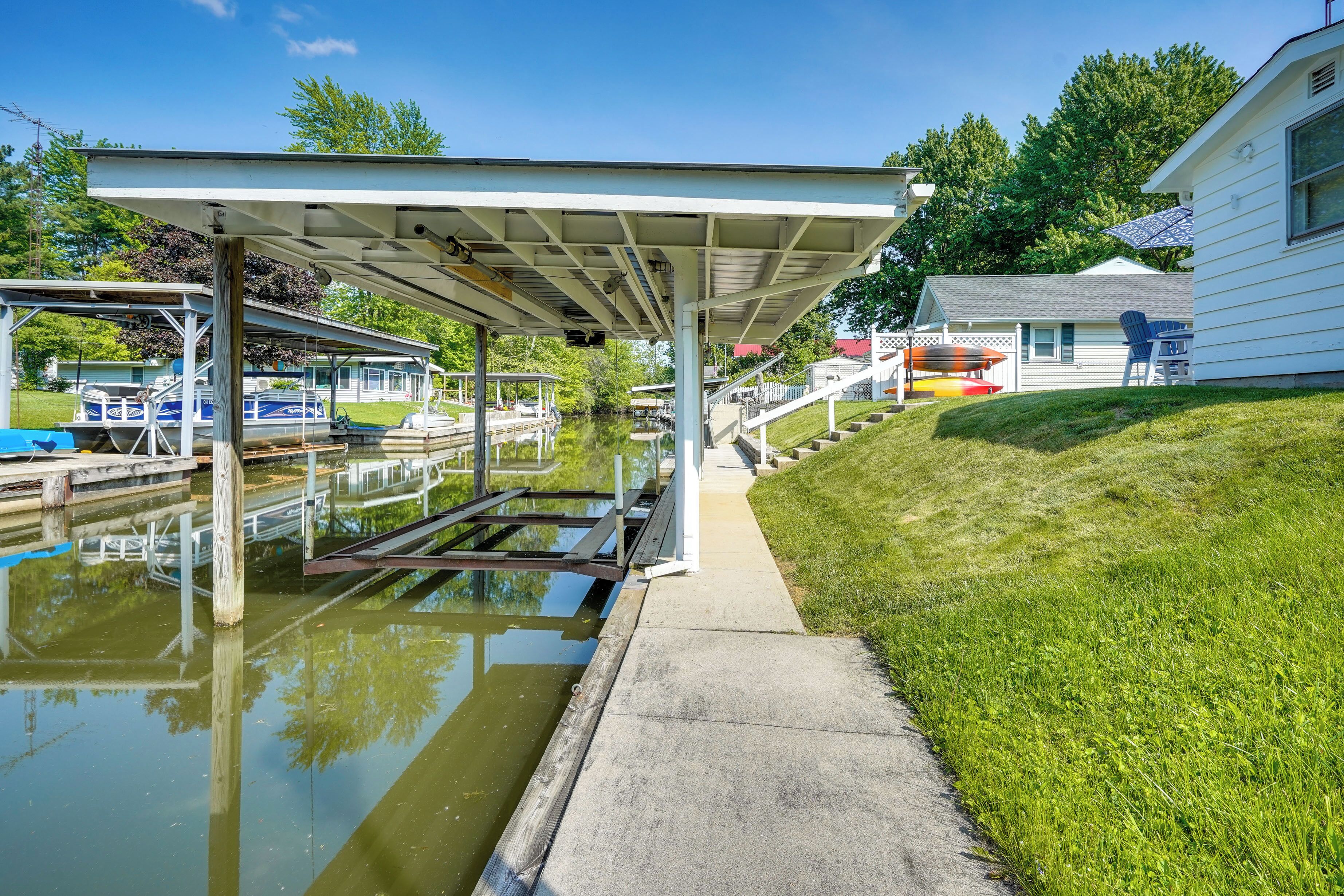 A covered dock area with a concrete walkway beside a calm water body, surrounded by greenery and a small house.