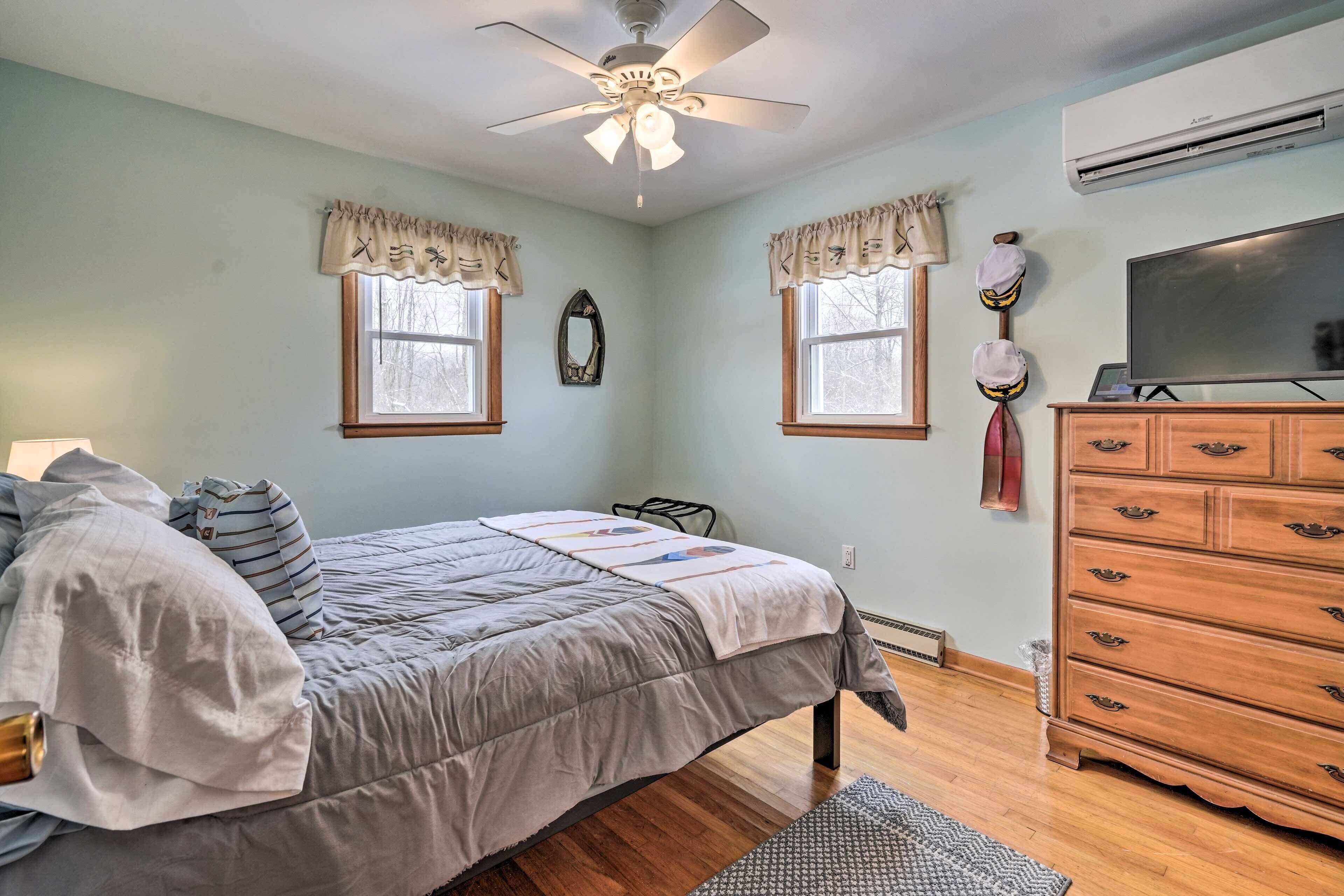 A bedroom with a bed, dresser, ceiling fan, two windows with curtains, and an air conditioning unit.