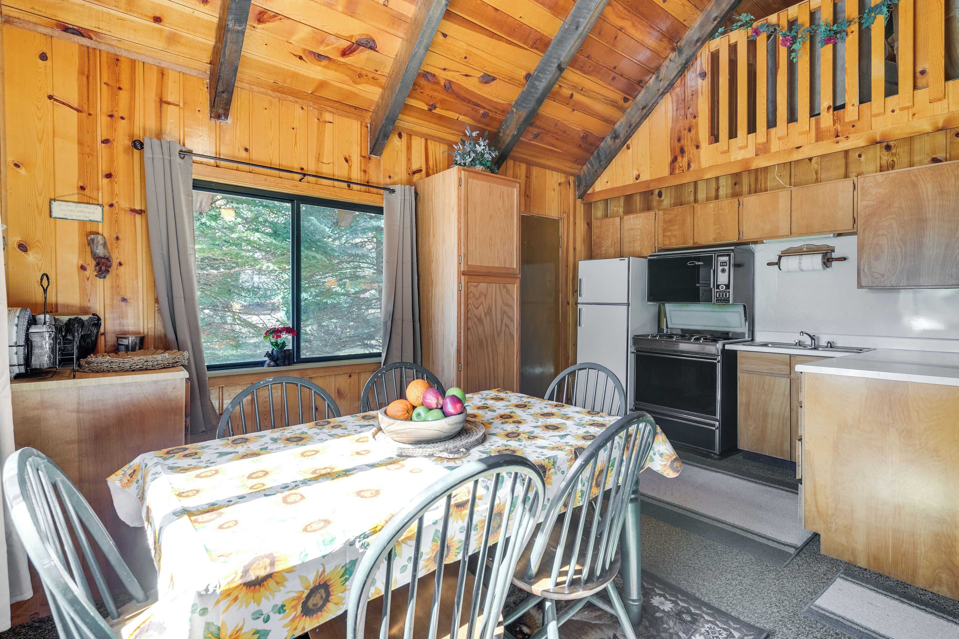 A wooden cabin interior with a dining table set for four, a kitchen with modern appliances, and a window overlooking greenery.