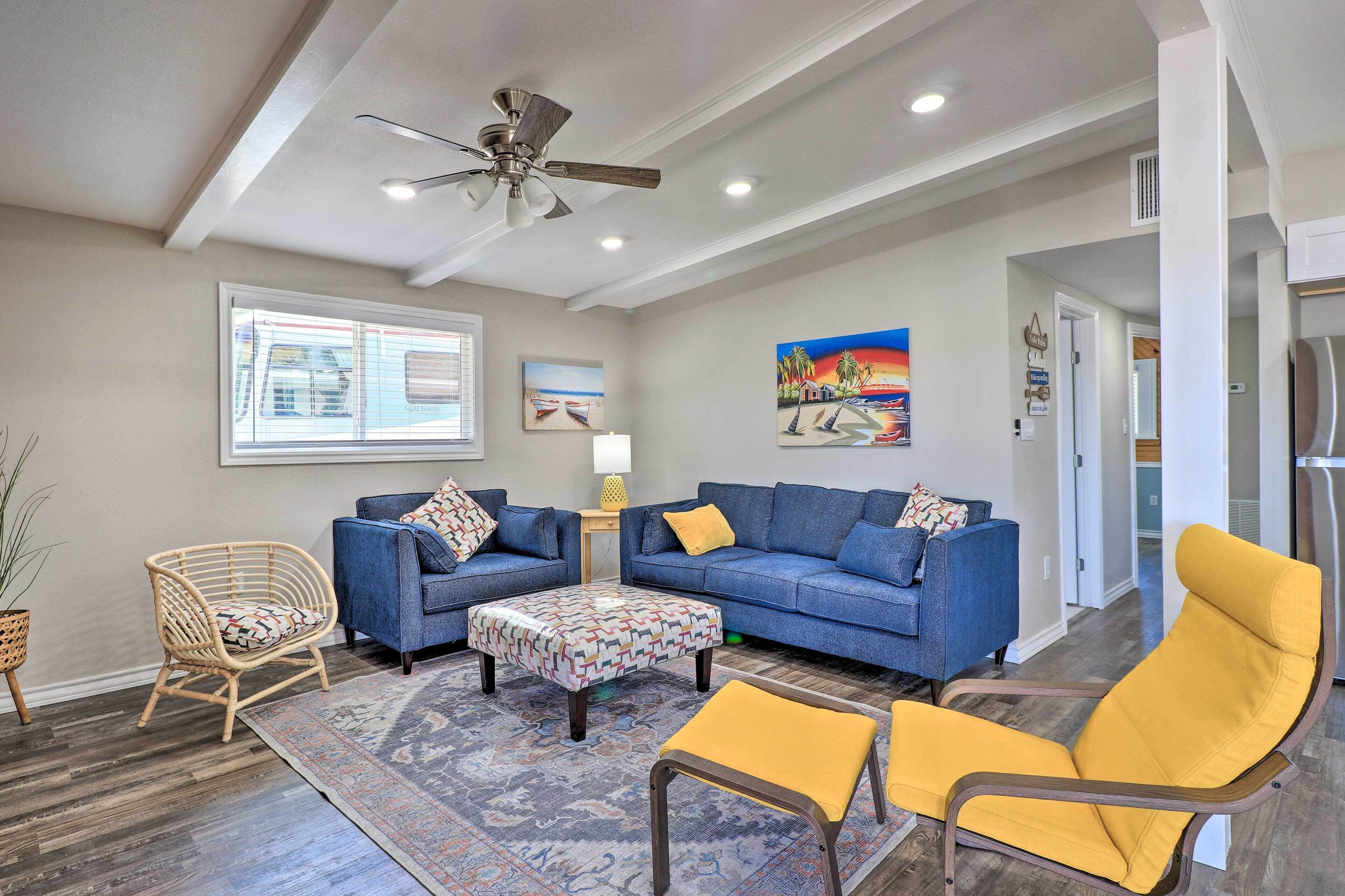 A living room with a blue sofa, a yellow armchair, a wicker chair, and a patterned ottoman.