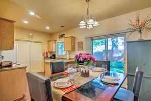 A dining area with a glass-top table, chairs, and a vase of flowers. There is a kitchen in the background with wooden cabinets and a clock on the wall. - Kanab Condo w/ Pool & AC < 1 Mi to Attractions! (Kanab)