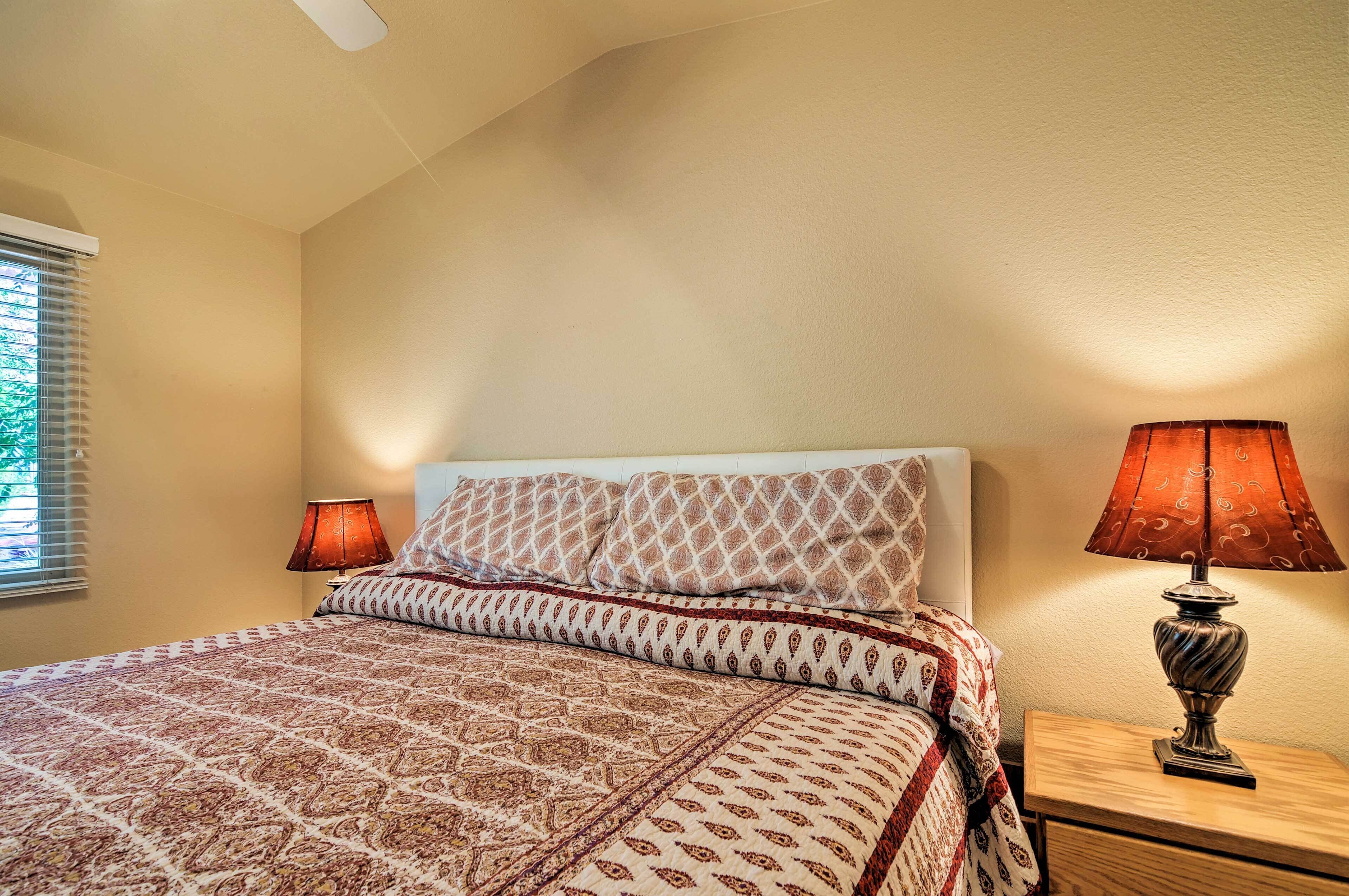 A bedroom with a bed, patterned bedding, two bedside lamps, a nightstand, and a window with blinds.