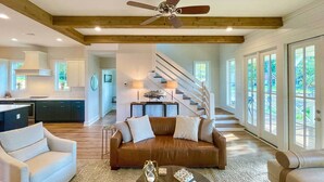 A spacious living area with a brown leather sofa, a white armchair, and a coffee table. A ceiling fan is visible above, and there is a staircase leading to another level. - Luxe Lakefront Getaway w/ Porch + Water View! (Dadeville)