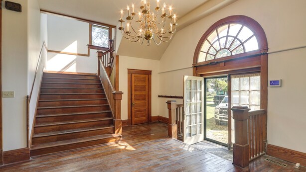 A spacious interior with a wooden staircase, a chandelier, and a large arched window.