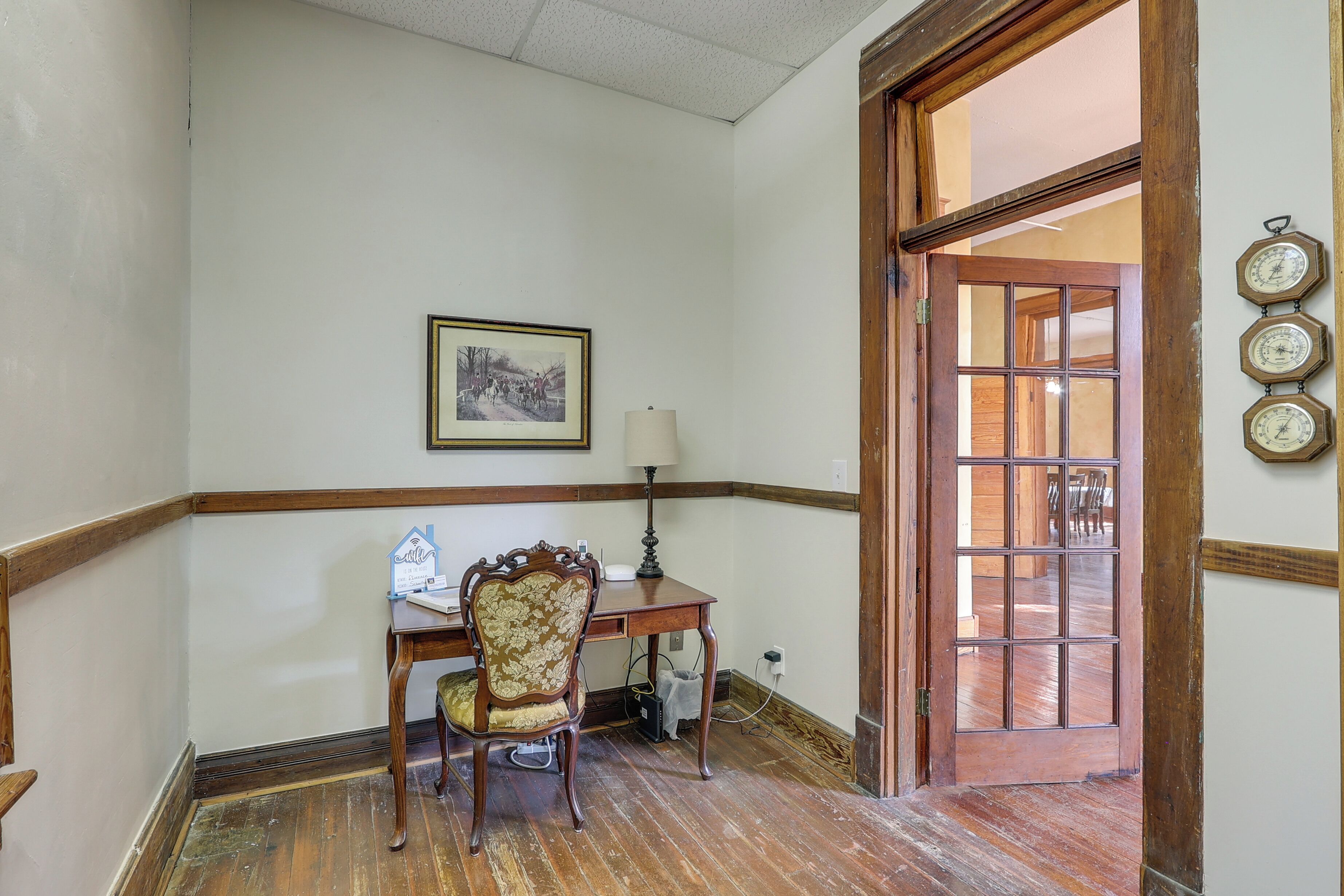 A room with a wooden desk, a chair, a framed picture on the wall, and a wooden door with a window.