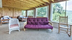 Une cabane avec un intérieur en bois, de grandes fenêtres, un canapé violet, une chaise en osier blanche et deux lits en bois.