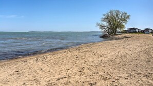 A sandy beach with a clear blue sky, a leafless tree, and a row of houses along the shore. - Home on Lake Erie 3 Bedrooms 3 Bathrooms (Port Clinton)