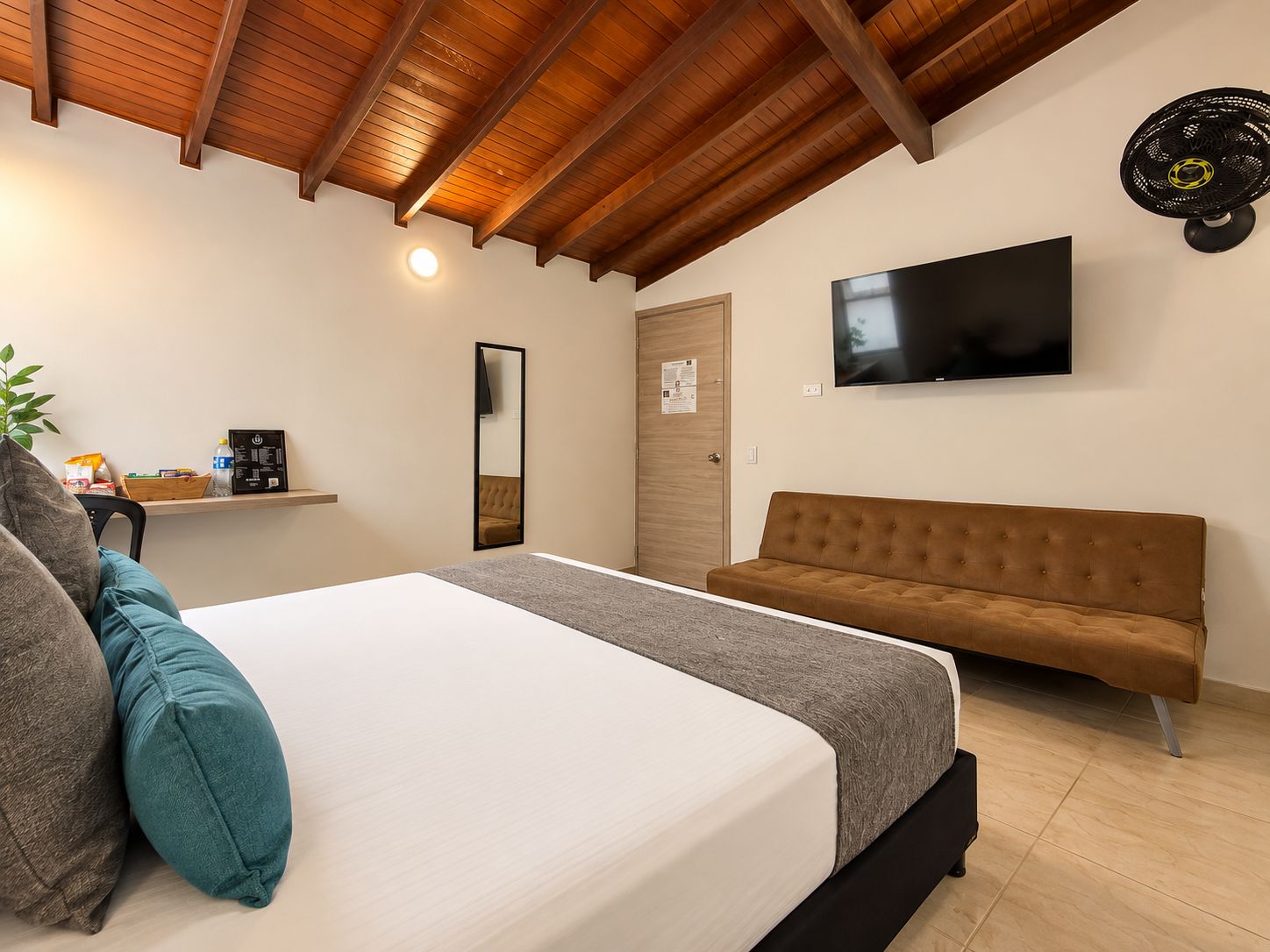 Una habitación de hotel con cama, sofá, televisión, espejo y repisa con objetos.