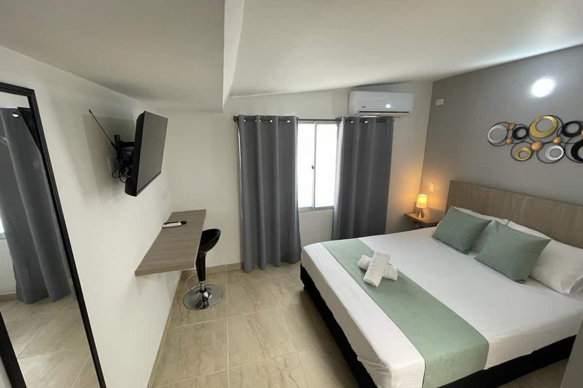 Quarto de hotel com uma cama, uma mesa, uma cadeira, uma televisão e ar condicionado fixado na parede.
