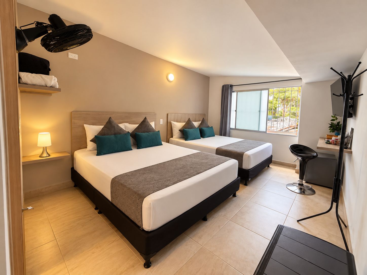 A hotel room with two beds, a desk, a chair, a standing fan, a lamp, and a window with a view.