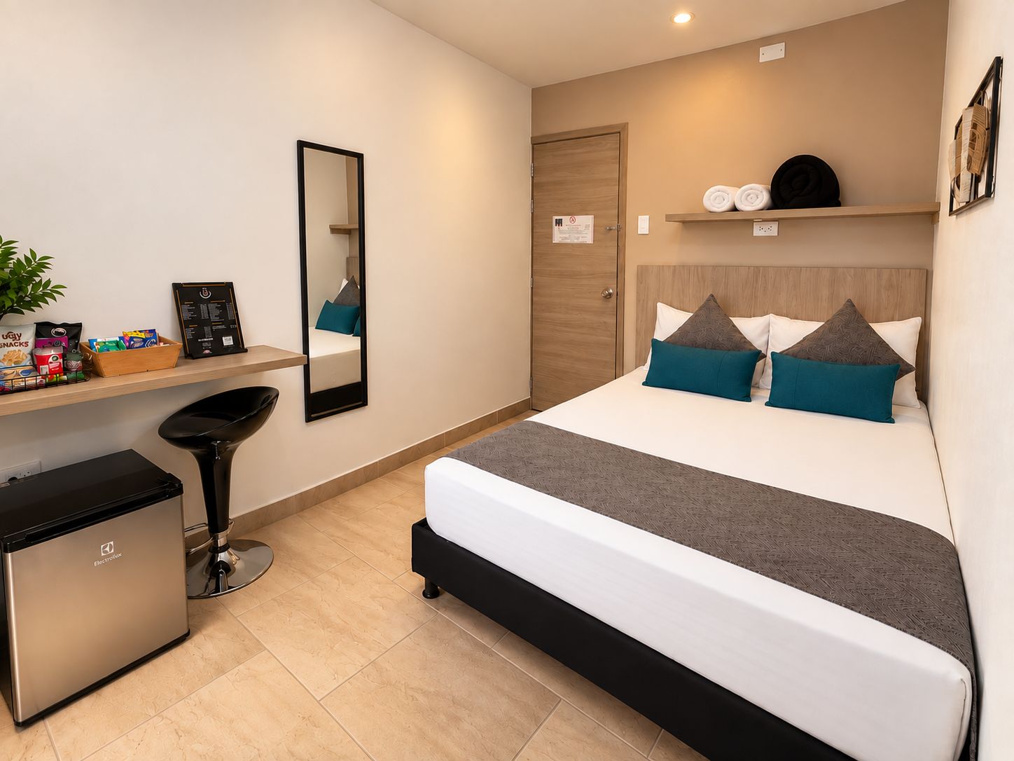 A modern hotel room with a bed, bedside table, mirror, and a small kitchenette.