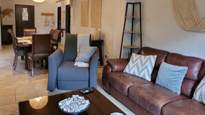 A living room with a brown leather sofa, a blue armchair, a coffee table with a bowl of seashells, and a dining area in the background. - 4th floor Apartment - 2 minutes walking distance to the beach! (Tamarindo)