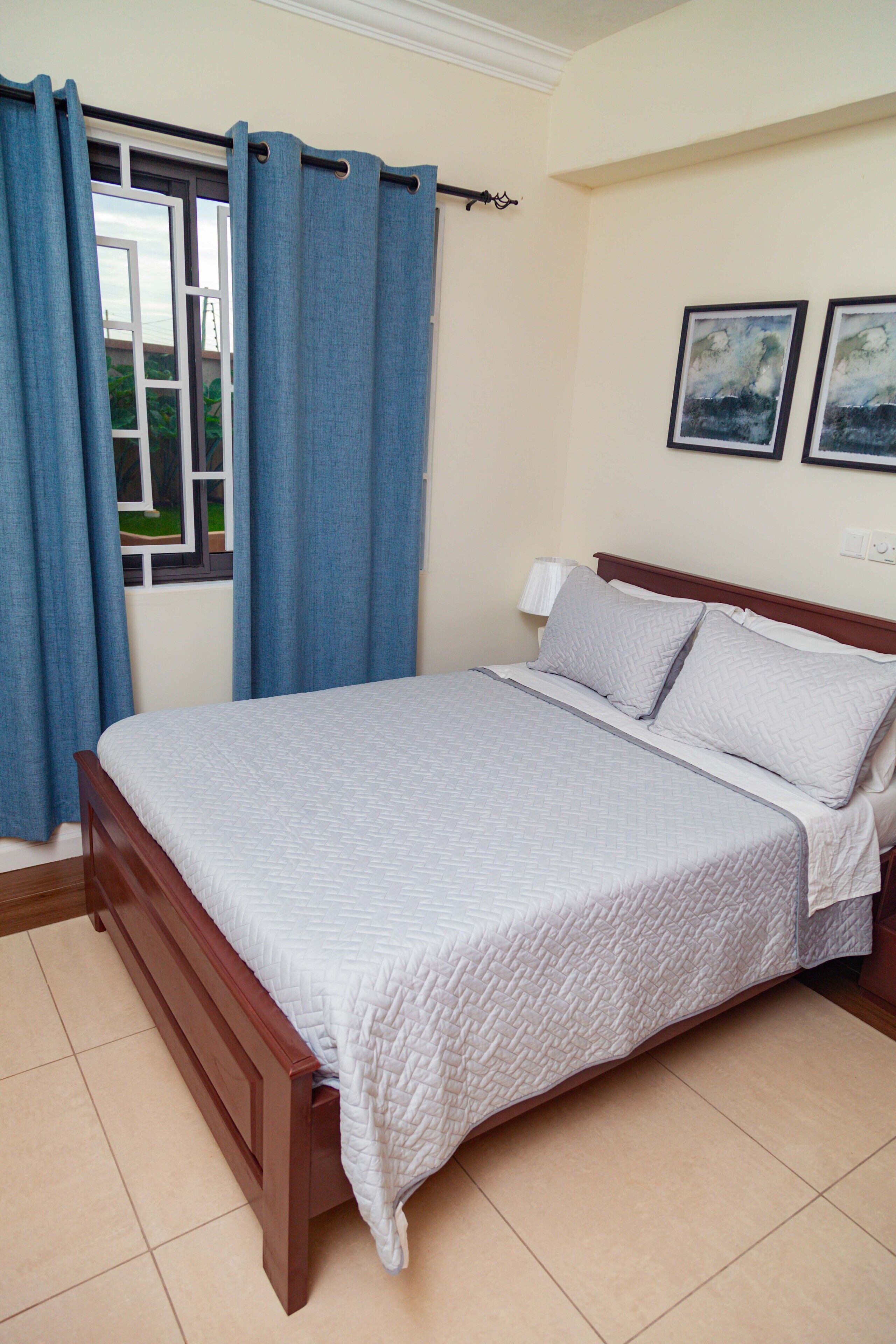 A bedroom with a wooden bed, a window with blue curtains, and two framed pictures on the wall.