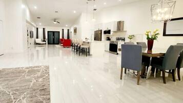 A spacious, modern living room with a marble floor, a dining area with a wooden table and chairs, and a kitchen area with white cabinets and stainless steel appliances.