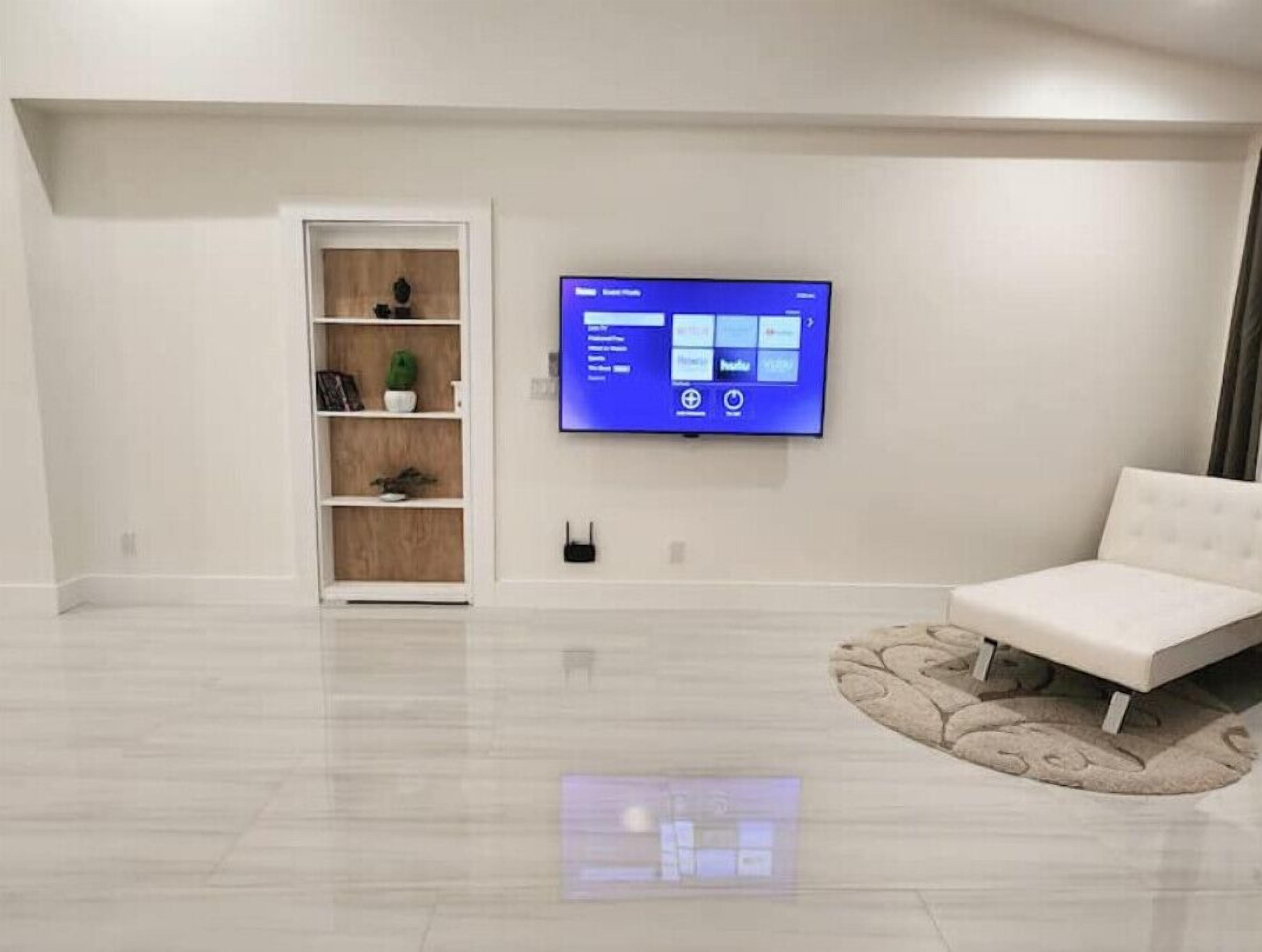 A modern living room with a flat-screen TV mounted on the wall, a white sofa, and a wooden shelving unit with decorative items.
