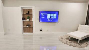 A modern living room with a flat-screen TV mounted on the wall, a white sofa, and a wooden shelving unit with decorative items.