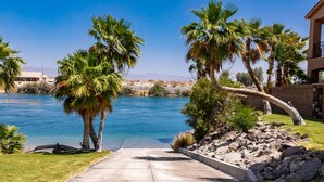 A paved pathway leading to a body of water, surrounded by palm trees and a building with a stone facade.