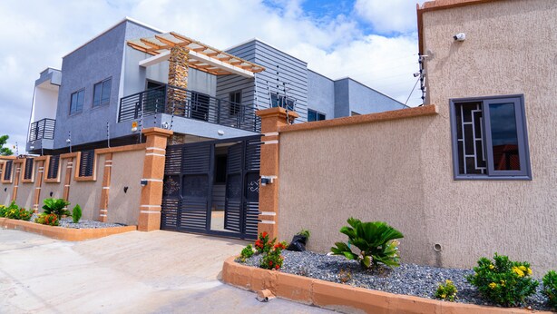 A modern two-story house with a gated entrance, a covered patio, and a small garden area. - Modern 2-Bedroom Apartment Home on a Gated Compound with Backup Solar Power. (Amrahia)