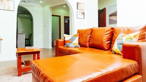 A modern living room with a leather sofa, a wooden coffee table, and a small side table. There are framed pictures on the wall and a doorway leading to another room. - Modern 2-Bedroom Apartment Home on a Gated Compound with Backup Solar Power. (Amrahia)