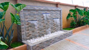 A rectangular water feature with multiple streams, surrounded by planters with large-leafed plants. - Modern 2-Bedroom Apartment Home on a Gated Compound with Backup Solar Power. (Amrahia)