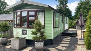 A small, single-story house with a green exterior and a brown roof. It features a large bay window on the front, a small porch, and a gravel driveway with stepping stones. - Gorgeous (sta)caravan in Tzummarum (Tzummarum)
