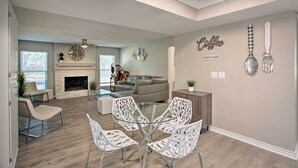A modern living room with a fireplace, a dining area with white chairs, and a kitchen area with a coffee-themed wall decoration. - Richardson Home w/ Pool, Hot Tub & Arcade Games (Richardson)