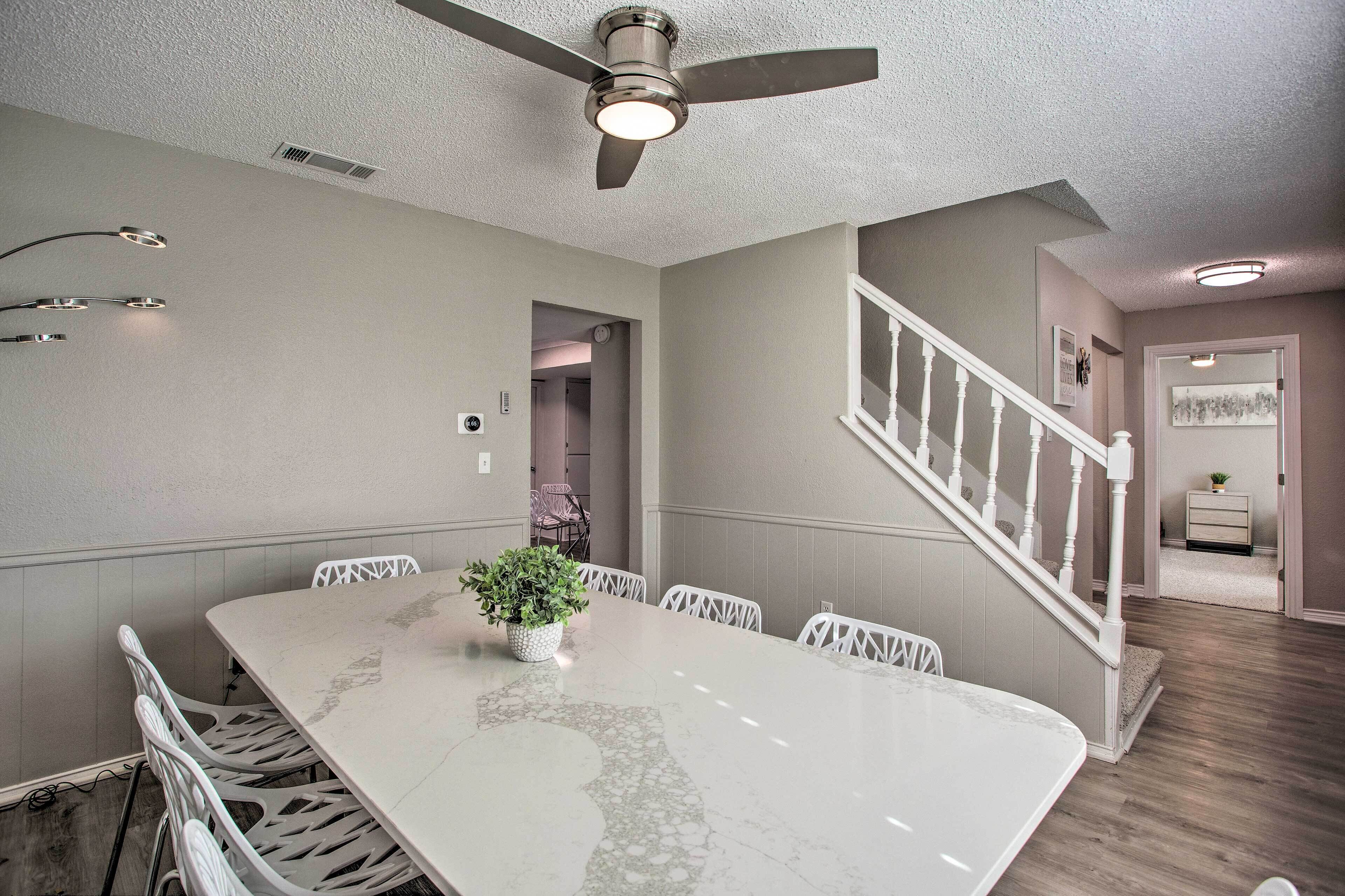 A dining area with a marble table, white chairs, a ceiling fan, and a staircase.