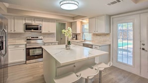 A modern kitchen with white cabinetry, stainless steel appliances, and a marble island with two bar stools. - Richardson Home w/ Pool, Hot Tub & Arcade Games (Richardson)