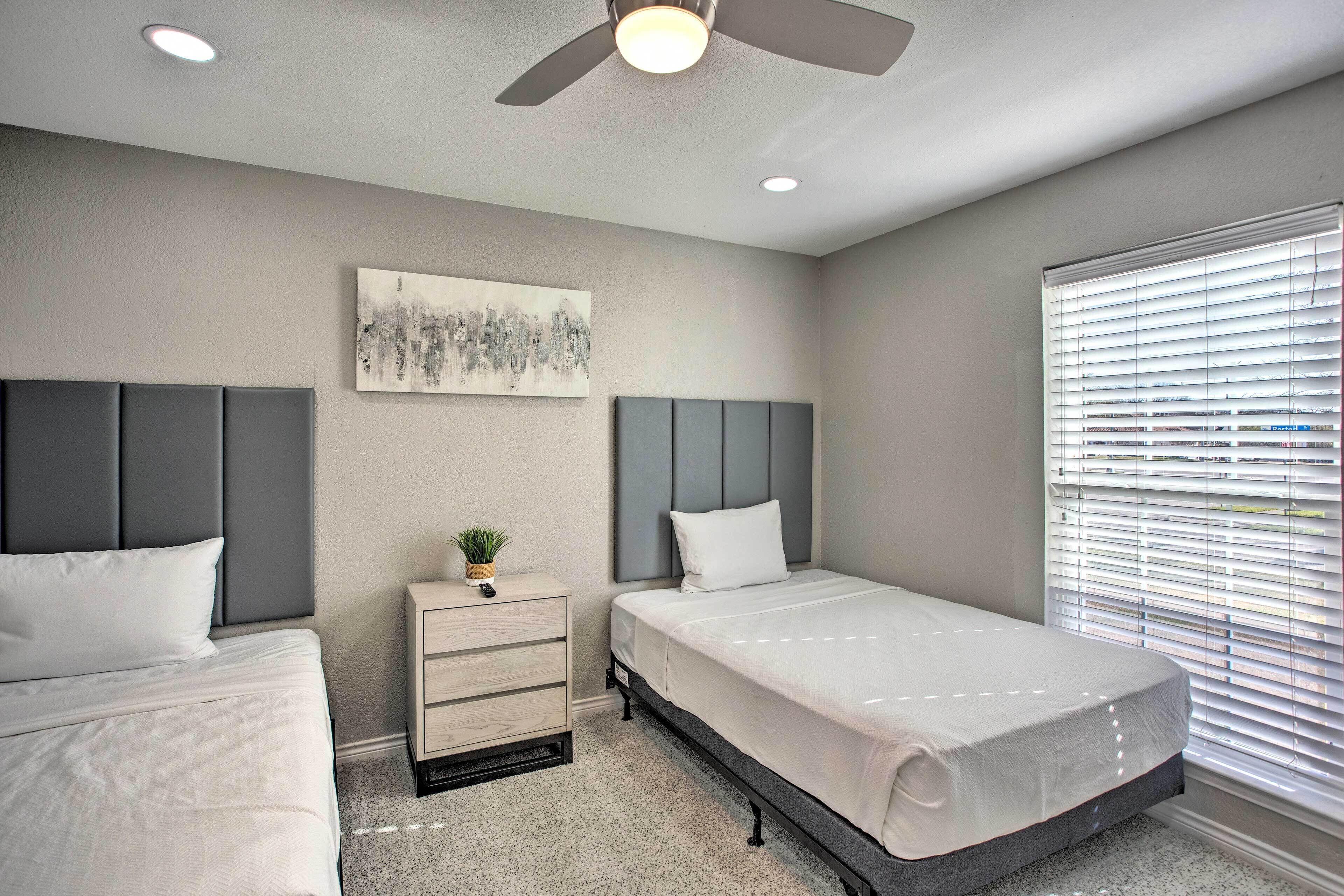 A bedroom with two beds, a nightstand, a ceiling fan, a window with blinds, and a wall-mounted artwork.