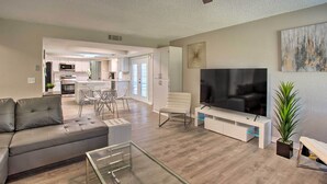 A modern living room with a grey sofa, a glass coffee table, a white TV stand, and a large flat-screen TV. - Richardson Home w/ Pool, Hot Tub & Arcade Games (Richardson)