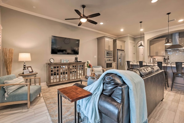 A modern living room with a sofa, a coffee table, and a TV. There is a kitchen area with cabinets and a refrigerator.