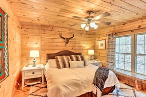 A cozy log cabin bedroom with a bed, two bedside tables, a ceiling fan, and a large window with a view of trees. - Deck & Game Room: Blue Ridge Mountain Cabin! (Warne)