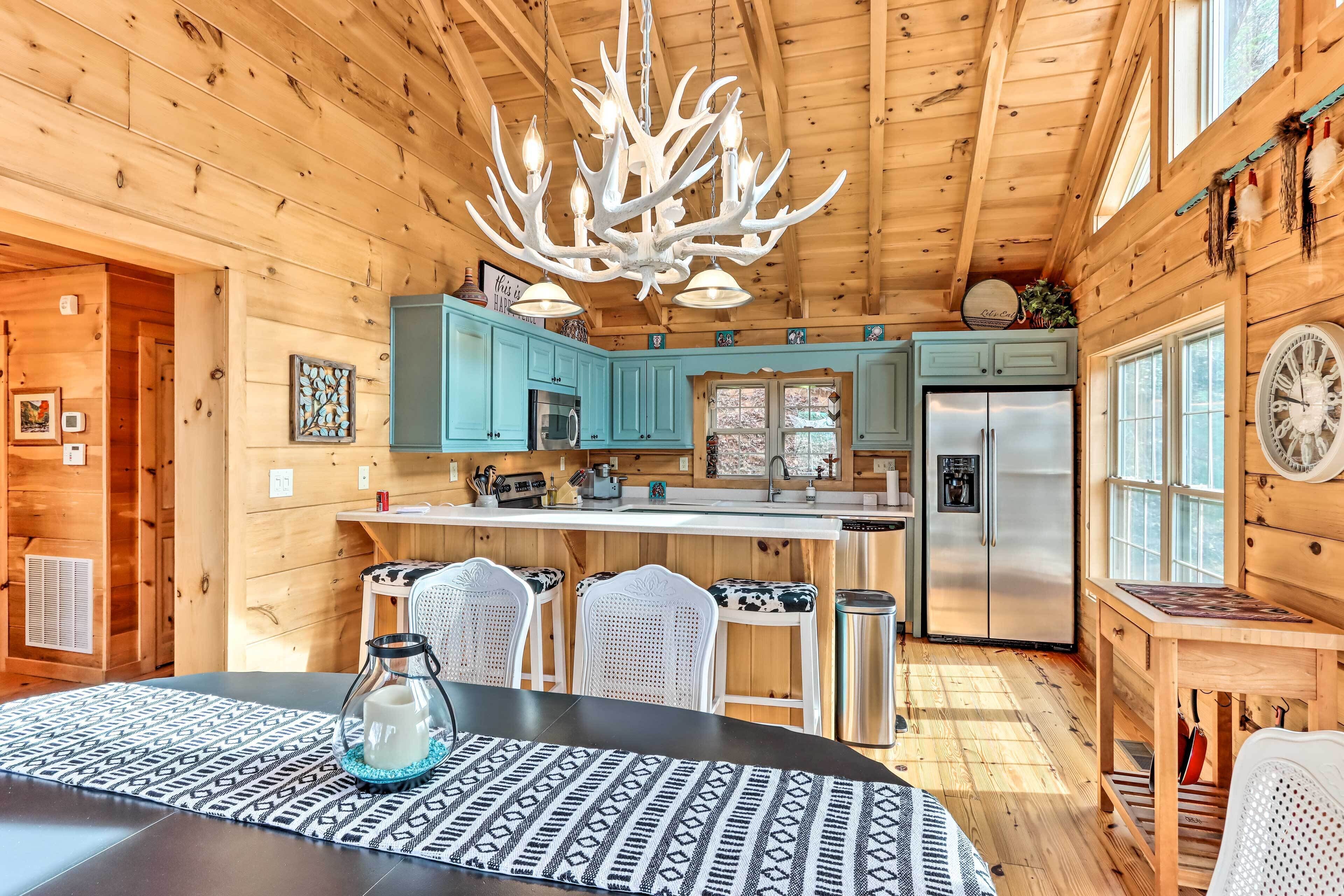 A wooden cabin interior with a kitchen, dining area, and a chandelier with antler decorations.