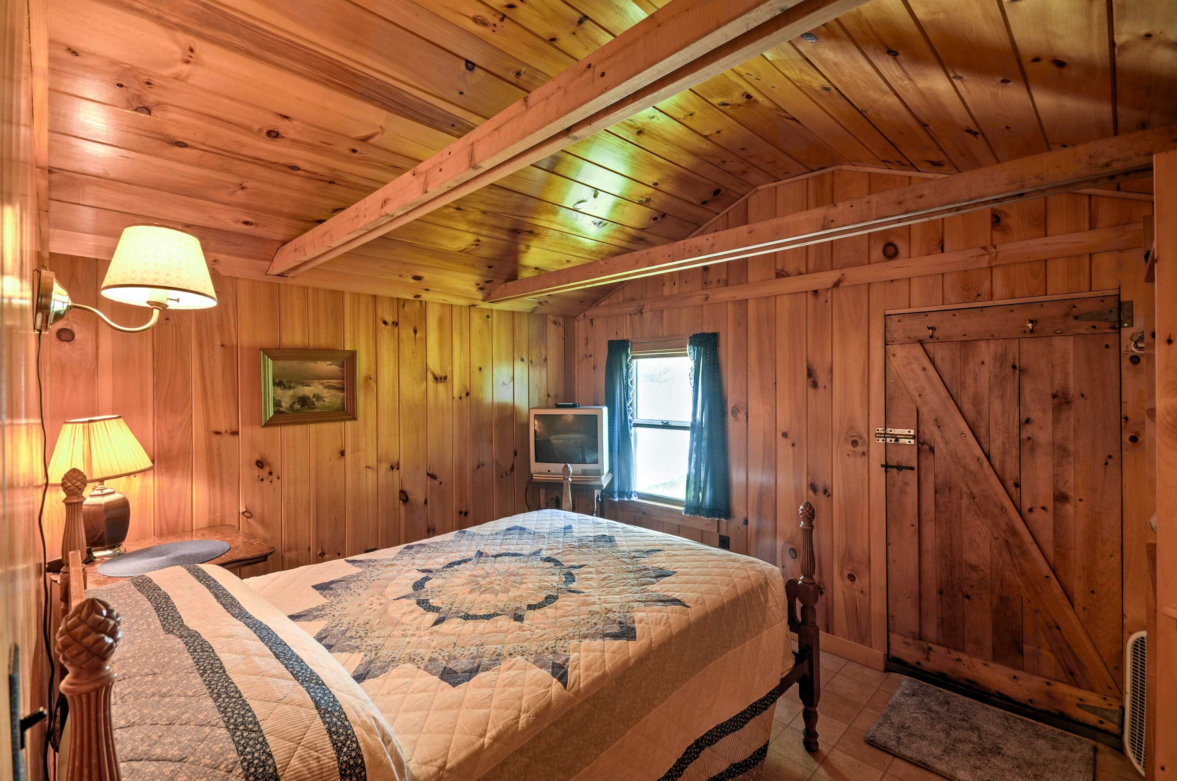 A wooden cabin bedroom with a bed, a television, a window with curtains, and a door.