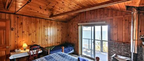 A cabin with wooden walls and ceiling, a bed with a quilt, a desk with a lamp, a chair, and a large window with a view of the outdoors.