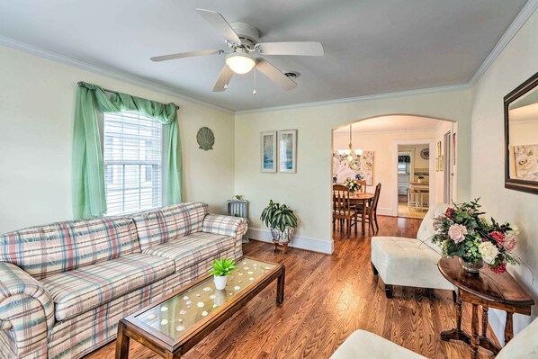 A living room with a plaid sofa, a glass coffee table, a white armchair, and a dining area in the background.