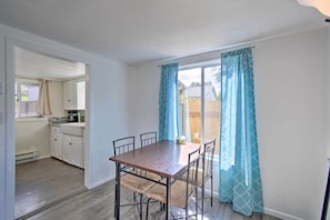 Een eetgedeelte met een houten tafel en stoelen, een keuken op de achtergrond en uitzicht op een achtertuin door een raam met blauwe gordijnen.