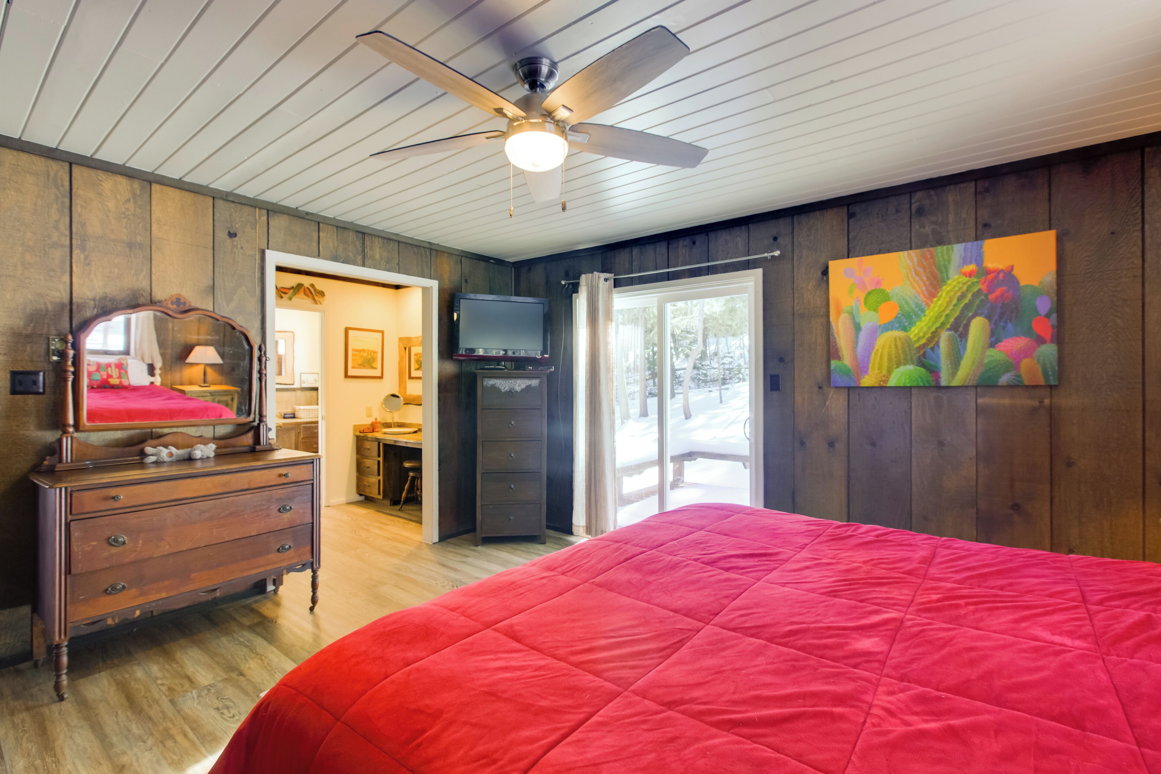 A bedroom with a large bed, wooden walls, a ceiling fan, a TV, and a sliding glass door to the outside.