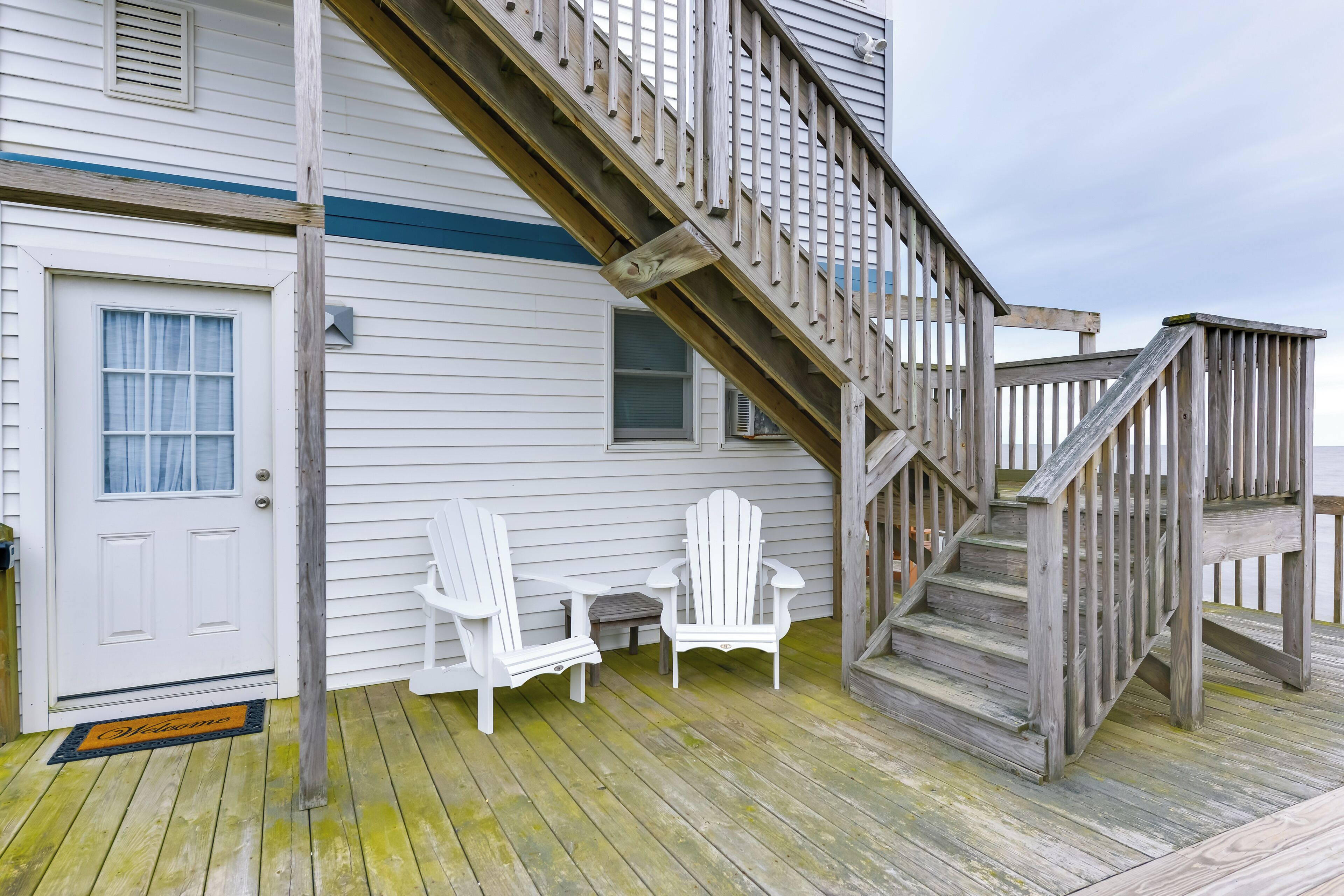 A wooden deck with a staircase leading to a second floor, white Adirondack chairs, and a small table.