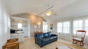 A spacious living area with a blue leather sofa, wooden rocking chair, and a dining table with chairs. A ceiling fan and pendant lights are visible. - Private Beach Access! Bayfront Cape May Cottage (Cape May Court House)
